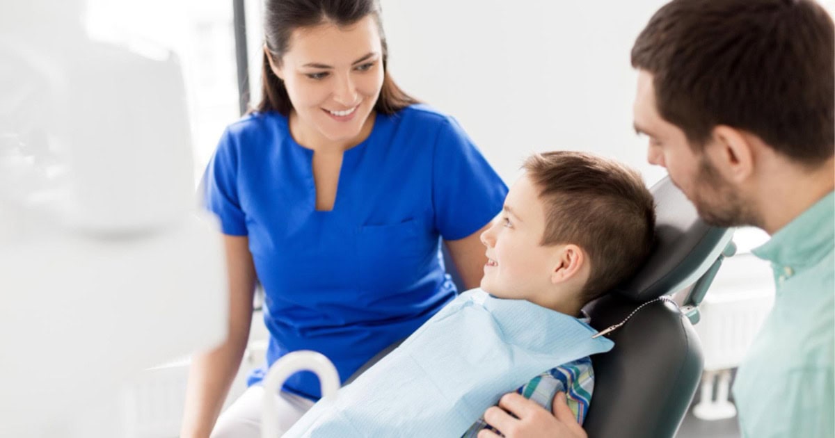 Boy smiling in a dental chair with his father beside him and a dentist looking on